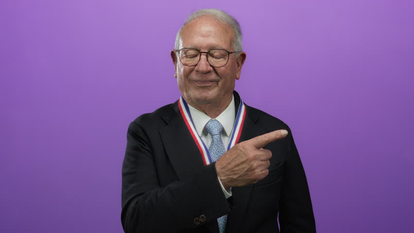 Senior man in business suit with silver hair wearing a medal, confidently pointing sideways against a vibrant purple wall background.