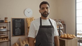 Young man crosses arms in bakery shop wearing apron surrounded by bread and pastries with clock and chalkboard in background - Powered by Shutterstock - Get 15% off with code: PIKWIZARD15