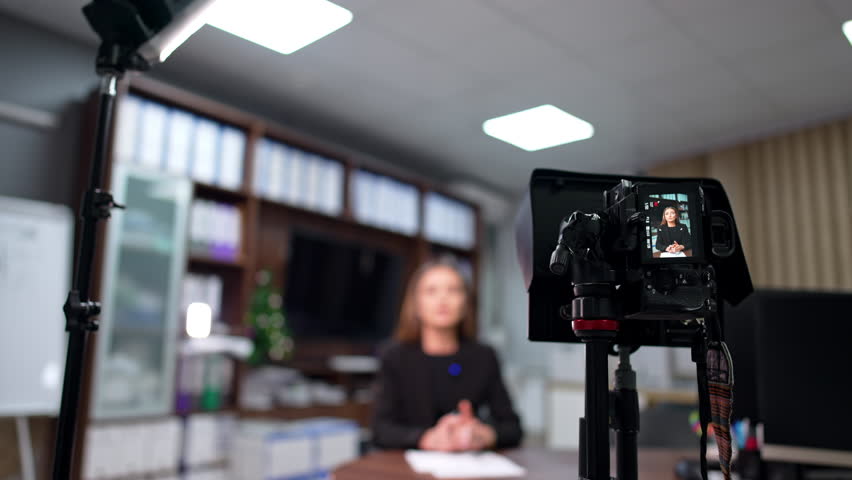 Modern camera on tripod shooting a blog content. Female speaker influencer sits at desk talking. Blurred backdrop.