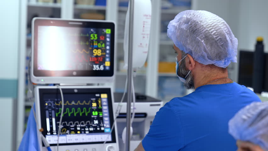 Rear view of the bearded doctor looking attentively at the monitors of lung ventilating machine. Anesthesiologist controls the condition of patient under surgery.