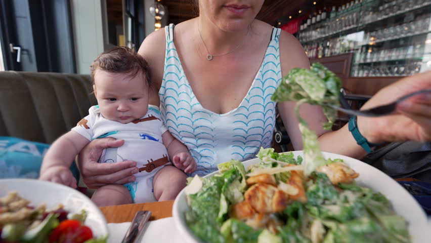 Baby sitting on mother’s lap while she eats a salad at a restaurant, showing a tender and nurturing family moment during a casual dining experience
