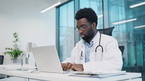 African American male doctor gesturing while sitting at desk, wearing stethoscope and glasses at laptop in bright office. Physician tapping and texting in keyboard during remote telehealth session. - Powered by Shutterstock - Get 15% off with code: PIKWIZARD15