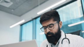 Close up of Indian male doctor in white coat with stethoscope, focusing on laptop, wearing glasses, sitting in modern office. Concentrated physician reviewing patient records on computer screen. - Powered by Shutterstock - Get 15% off with code: PIKWIZARD15