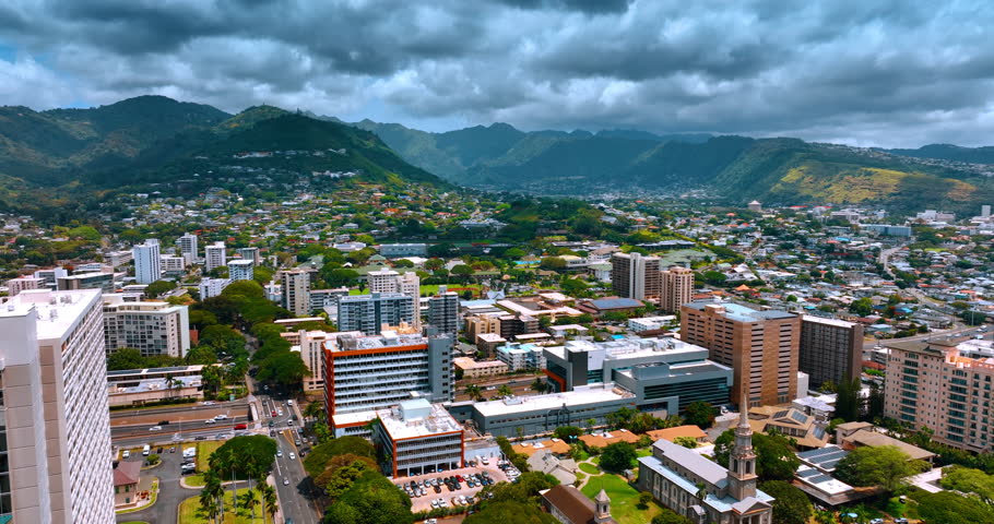 Approaching verdant mountains surrounding the vast city. Dramatic cloudscape covers the sky over beautiful Honolulu, Hawaii, USA. Aerial view.