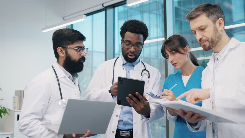 Multiracial team of doctors and nurses, standing close and discussing diagnosis, reviewing notes and tablet, wearing white lab coats and scrubs, in modern office. Diverse healthcare professionals. - Powered by Shutterstock - Get 15% off with code: PIKWIZARD15