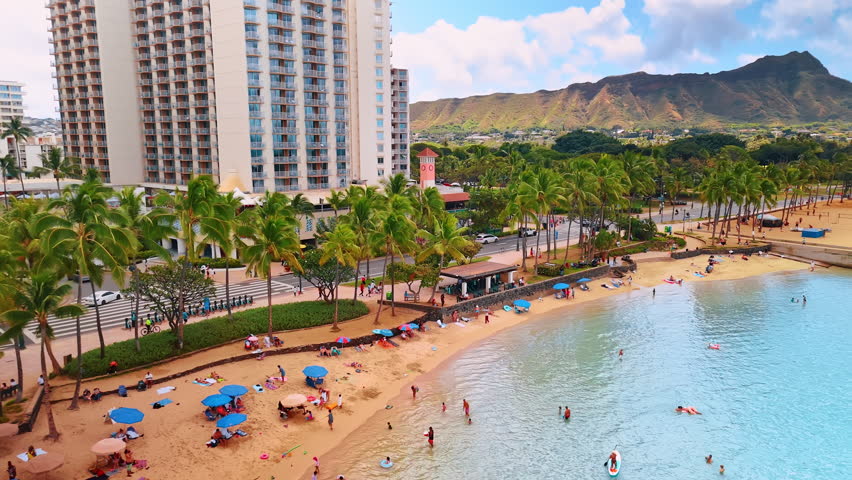 Flight along the sandy beach with green palm trees on. Shore of Waikiki with vacationers resting. Diamond Head Crater at backdrop. Honolulu, USA.