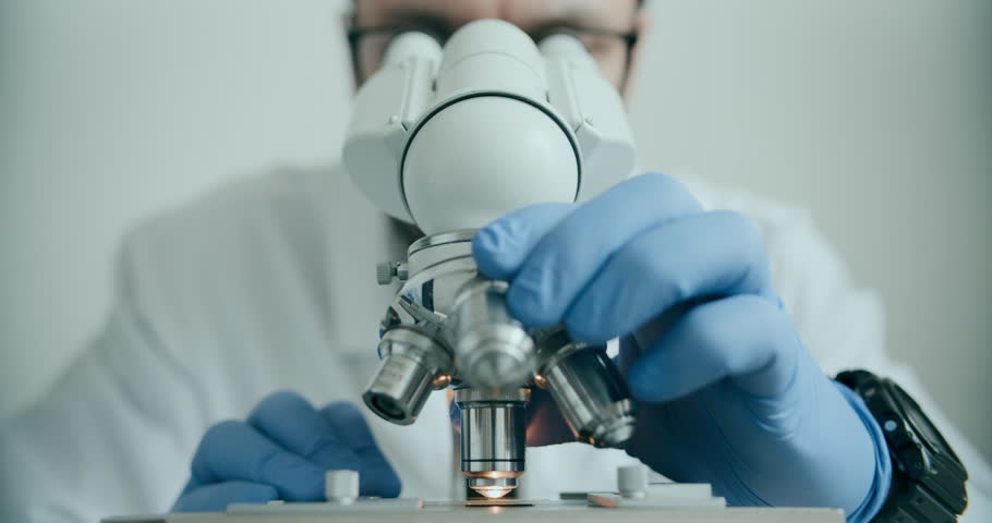 Close-up of biologist gloved hands using microscope in sterile laboratory. Vibrant blue gloves highlight precise scientific work against monochromatic lab backdrop