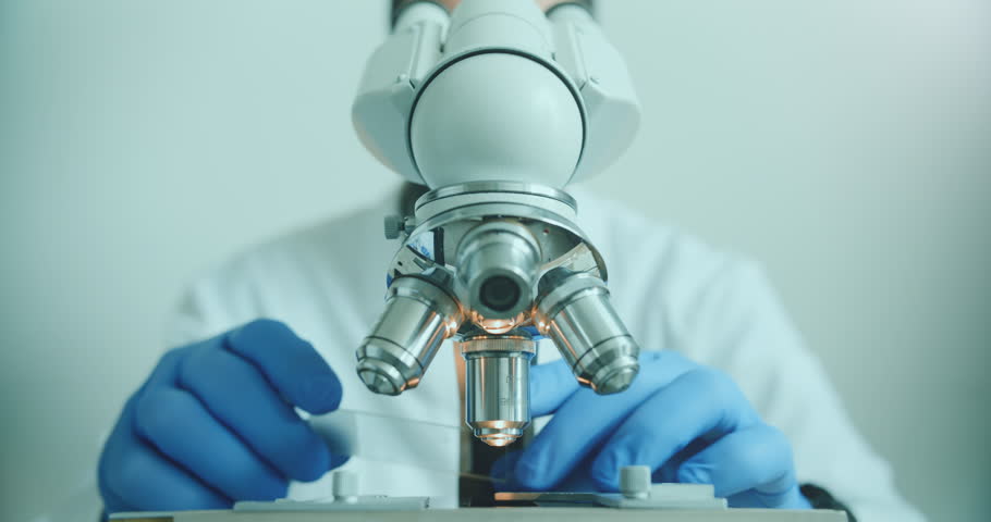 Close-up of biologist gloved hands using microscope in sterile laboratory. Vibrant blue gloves highlight precise scientific work against monochromatic lab backdrop