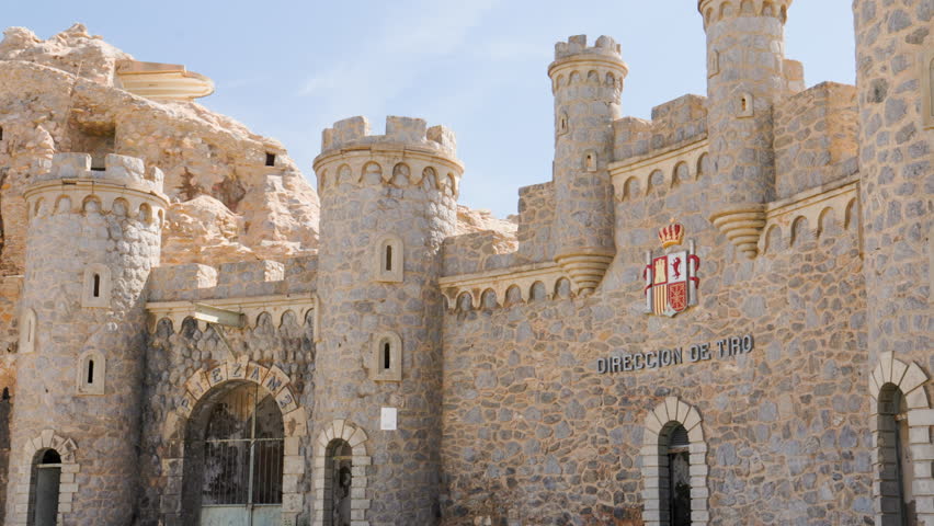Front view of Batería de Castillitos fortress in Cartagena, Spain, showing detailed stone towers, Spanish coat of arms, and arched military entryways under clear blue sky on rocky hillside