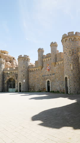 Vertical view of Batería de Castillitos fortress facade in Cartagena, Spain, with medieval-style stone towers, Spanish coat of arms, and bright daylight casting strong shadows on paved ground
