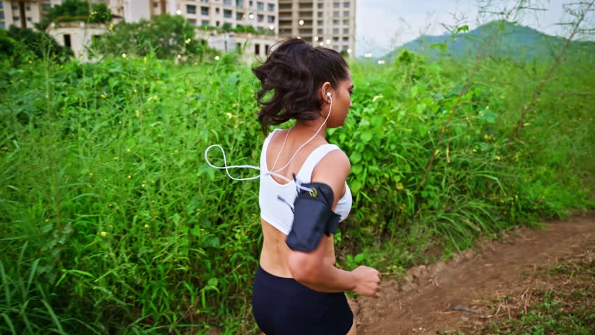 Female follows dirt path wearing sports bra and shorts, Woman Jogging with Smartphone Armband Outdoors, Lady devotee jogs track in retreat, Jogger explores lush terrain for wellness, Daily routine