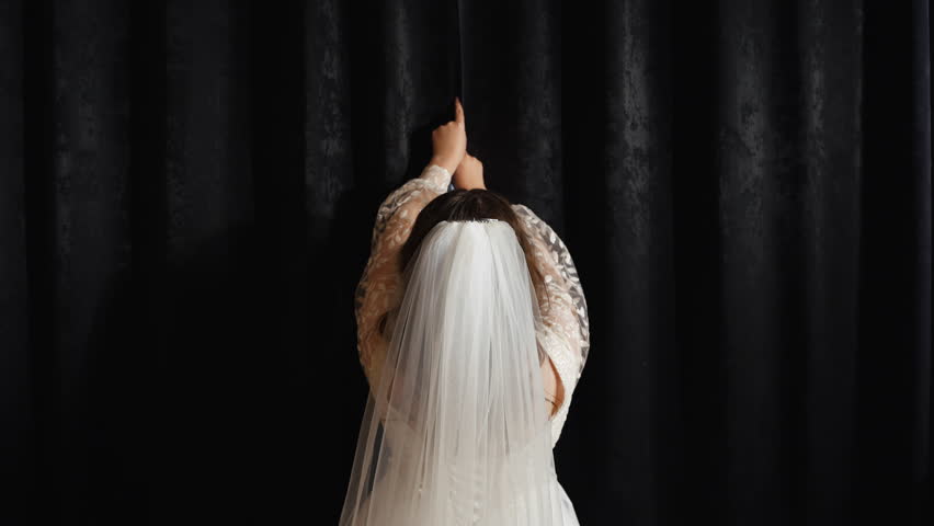Back view of a bride in a white dress and with veil on the head, opening black curtains to the sides in front of a window with her female hands in room. Symbolic wedding tradition. No face