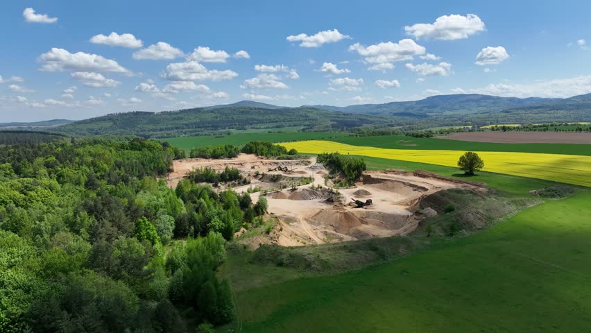 Aerial drone view of sand or gravel quarry surrounded by forest and farmland with distant hills. Aerial Drone view shows an inactive or abandoned quarry embedded in a green landscape.