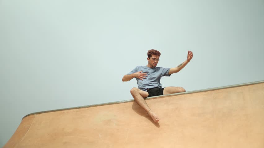 Young male casually sitting on wall, making diverse hand and body gestures while wearing casual summer outfit against minimal urban background