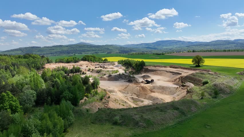 Aerial drone view of sand or gravel quarry surrounded by forest and farmland with distant hills. Aerial Drone view shows an inactive or abandoned quarry embedded in a green landscape.