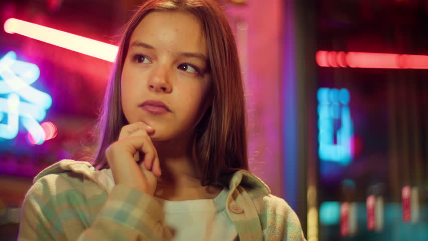 Puzzled Caucasian Asian junior kid contemplate in city schoolgirl brainstorm girl child think about problem teen teenage pensive evening neon. The inscription noodle in Chinese. Translation: 'noodle'.