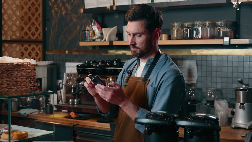 Happy Caucasian barista worker waiter guy scroll screen mobile phone employee male laughing in cafeteria bartender man typing smartphone cyberspace online small business Internet shopping social media