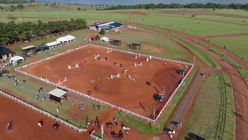 Overhead aerial orbit of horse arena with red sand ring and stables around its perimeter, competitors exhibition practice