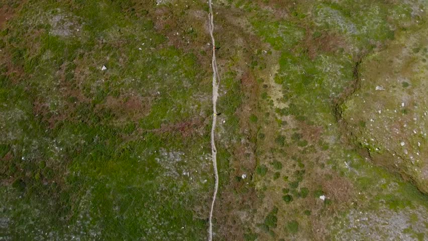 Top down aerial view of a lonely mud hiking road or pathway going straight across a grassy green and brown Sweden landscape into the distance during a cloudy day. The ground has big rocks and moss.