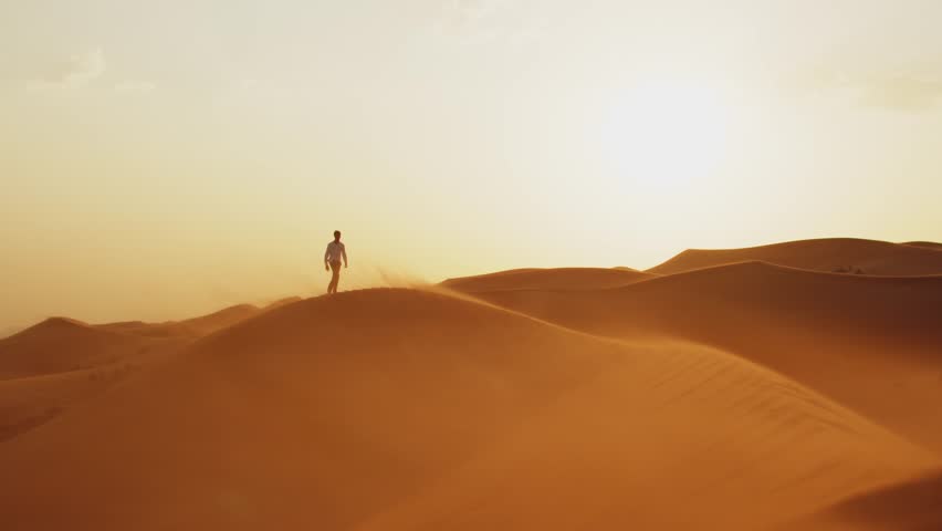 Slow motion video: Silhouette of a human walking on the sands in the desert in windy weather while sunrise.  Young man in stylish clothes walks barefoot on the sand dunes in the desert at sunset. 
