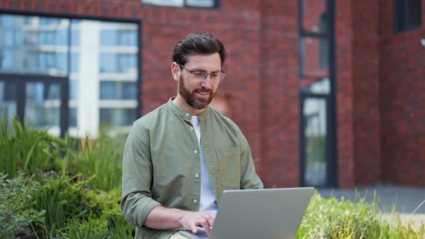 Handsome businessman working outdoors. Successful freelancer typing on laptop, sitting near office building. Professional specialist in casual clothes feels happy and inspired.