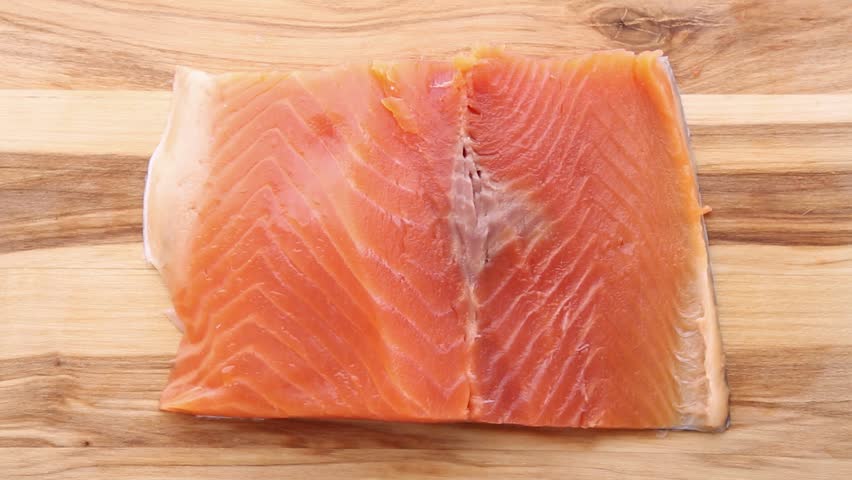 Fresh salmon fillet being cut open with sharp knife on wooden cutting board. Chef skillfully slices through the fish revealing inner flesh, then spreads it open with hands for preparation.