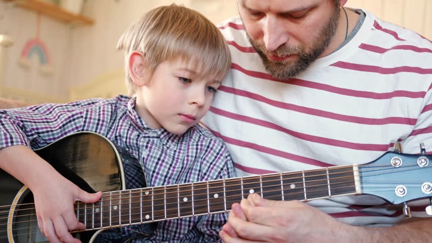 Handsome dad teaches his cute son to play the guitar sitting on sofa in the living room at home. Happy Father's Day. Father teaching his child to play guitar. Kid Learning music skill. Art education