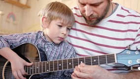 Handsome dad teaches his cute son to play the guitar sitting on sofa in the living room at home. Happy Father's Day. Father teaching his child to play guitar. Kid Learning music skill. Art education - Powered by Shutterstock - Get 15% off with code: PIKWIZARD15