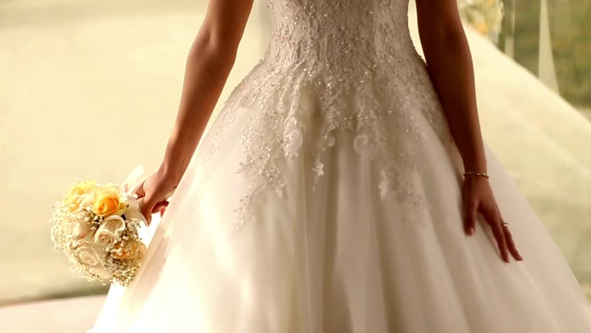 A bride stands outdoors, showcasing her stunning white gown and holding a vibrant bouquet of flowers. The setting is bright and inviting, capturing the joy of her special day.