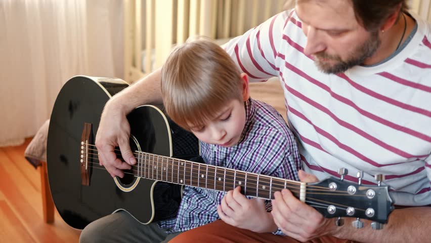 Handsome dad teaches his cute son to play the guitar sitting on sofa in the living room at home. Happy Father's Day. Father teaching his child to play guitar. Kid Learning music skill. Art education