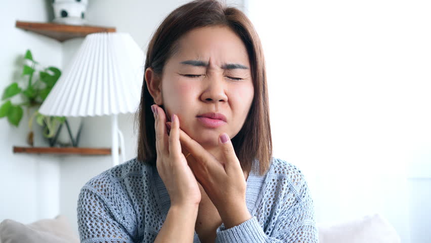 Asian woman in discomfort, touching her cheek due to a throbbing toothache and swollen face, likely caused by serious oral problems such as cavities, gum disease, or dental abscess