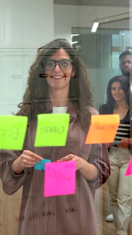 Businesswomen using colorful sticky notes on a glass wall during a brainstorming session, planning and organizing their work