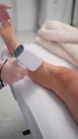 Laser hair removal on female leg in beauty salon, close-up of handheld device during procedure. Modern aesthetic treatment for smooth skin, performed by professional with protective gloves.