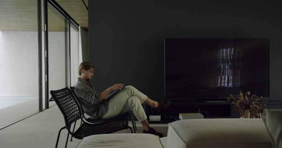A man relaxes in a wicker chair by a large window, immersed in a book within his sleek, modern living room. Man Relaxing in Modern Home with Book by Large Window