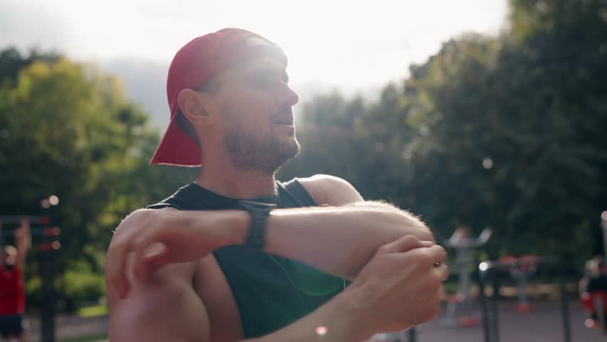 A young man stretches his arms in a park. He is wearing a black tank top and red baseball cap, preparing for a workout. Other people can be seen exercising in the background.