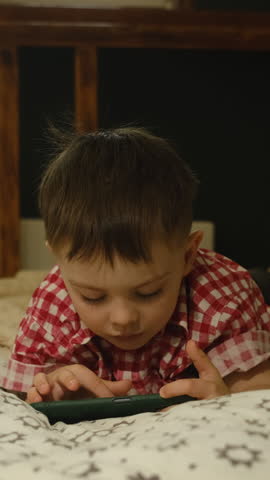 Vertical video. Preschool age child using mobile phone lying on bed at home 