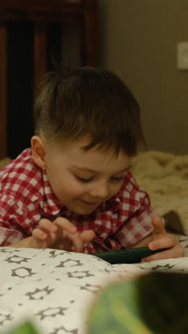 Vertical video. Child five years old lying on bed and using mobile phone. Home entertainment concept.