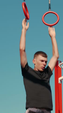 Male athlete exercising on gymnastic rings outdoors. Man practicing hard cardio endurance workout using gymnastic rings on sport ground. Vertical video