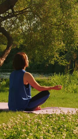 Woman doing yoga exercises in summer city park. Peaceful female meditating in the morning, enjoying sunrise. Girl practicing yoga, sitting in lotus position. Vertical video