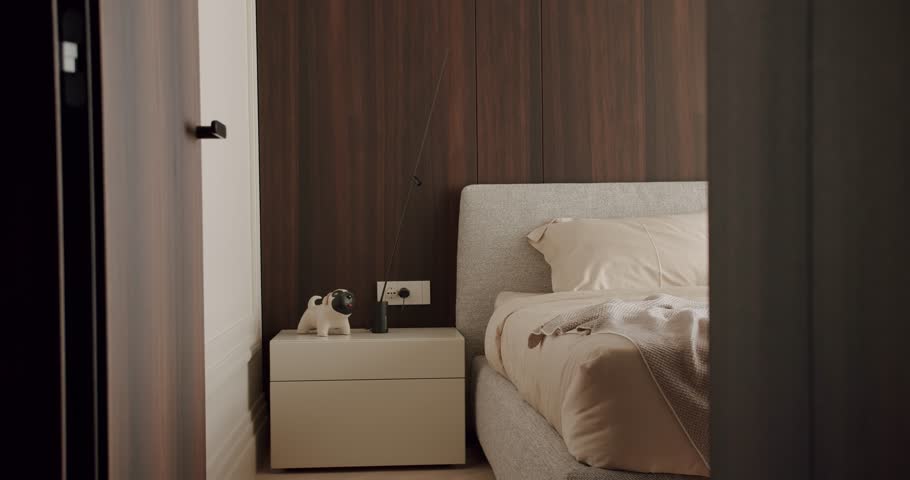 A modern and minimalist bedroom featuring a cozy bed with beige linens, a stylish nightstand with a small dog statue, and a dark wooden panel wall, creating a warm and inviting atmosphere.