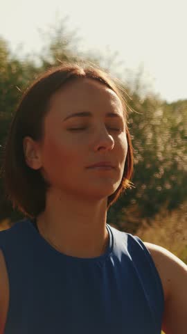 Portrait of calm young woman taking deep breath of fresh air, relaxing and meditating with closed eyes at sunrise. Female meditating in park, enjoying peace. Vertical video