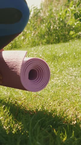 Female rolling out her exercising mat before yoga practice on green grass in park in the morning. Woman rolling out yoga mat and preparing to meditate training. Vertical video