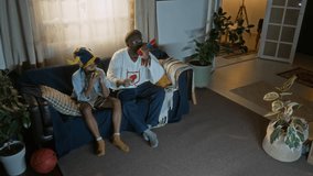 Cheerful African American father and son seated on couch, with snacks and noisemakers in hands, fully engaged in sports game night tradition from comfort of their living room - Powered by Shutterstock - Get 15% off with code: PIKWIZARD15