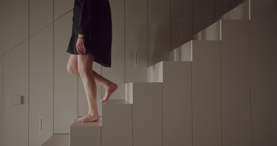 A woman in a black dress walks elegantly on a sequence of high, neutral-toned geometrically shaped cabinets in a modern, minimalist room. The image captures a sense of balance and minimalism.