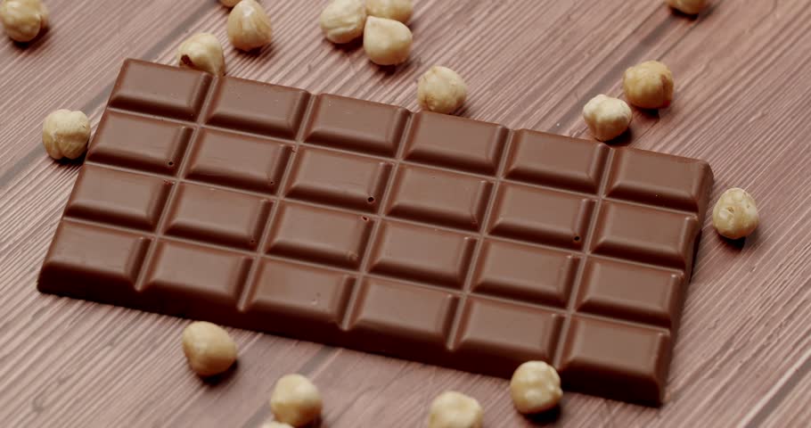 Close-up of a delicious milk chocolate bar surrounded by whole hazelnuts on a textured wooden surface. Perfect for depicting indulgence, sweetness, and gourmet treats.