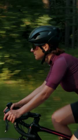 Young sportive woman rides on road bike, side follow shot. Fitness female athlete in sports clothes and helmet is cycling in park. Professional cyclist is training on bicycle. Vertical video