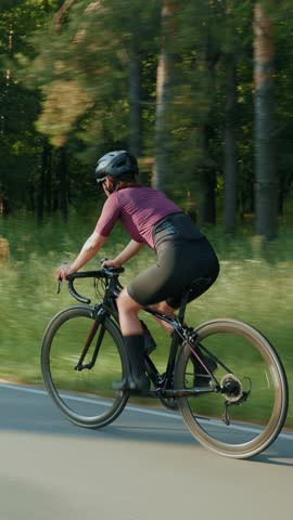 Female in cycling apparel and helmet pedaling hard out of saddle on road bike. Woman is cycling fast on bicycle in park. Professional sportswoman is training for triathlon competition. Vertical video