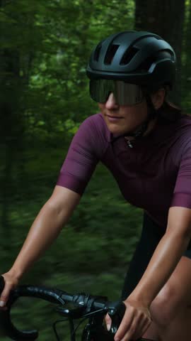 Portrait of woman in cycling apparel and helmet cycling bicycle on gravel forest road. Cyclist riding bike, travelling along green park. Vertical video