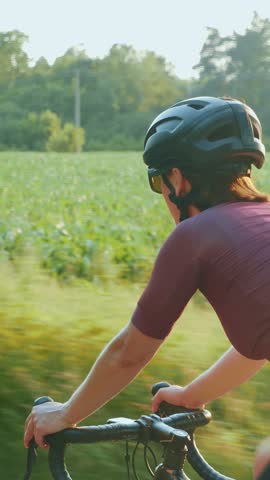 Cyclist is pedaling on cycle path in park at sunset. Woman in stylish cycling apparel and helmet riding on road bicycle. Vertical video