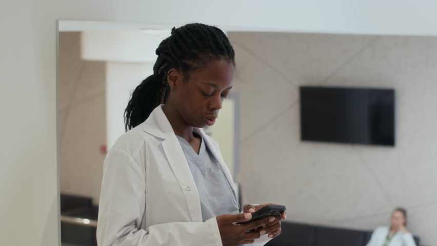Static shot of Black female healthcare worker text messaging on mobile phone then answering incoming call while standing in clinic hallway then walking off camera